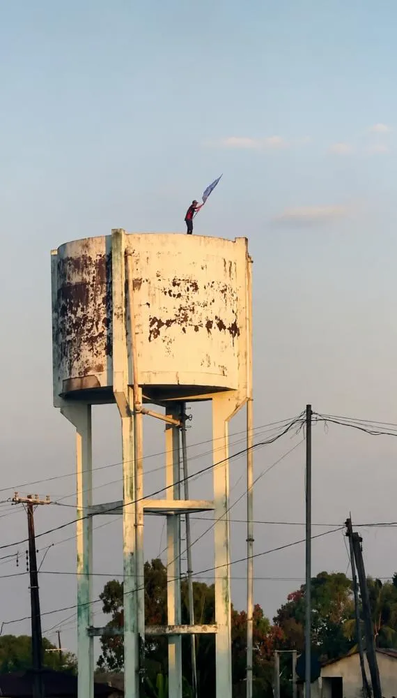Homem aparece sobre uma caixa d’água no bairro Cajue Acre, em Plácido de Castro, enquanto levanta uma bandeira de cunho político - Foto: Captura de Tela.